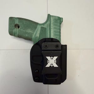 Op de afbeelding: Een saliegroen handvuurwapen dat gedeeltelijk in een zwarte, gevormde holster is geplaatst. De holster is voorzien van een zwarte riemclip met een wit gestileerd X-logo in het midden. De holster is bevestigd met twee zwarte schroeven aan de bovenkant.