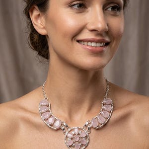 May include: A silver-toned necklace featuring a central circular pendant adorned with multiple teardrop-shaped pale pink stones, flanked by matching teardrop stones along the chain, all set in a metallic frame with a link chain closure.
