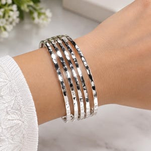 May include: A set of four silver-toned bangles featuring a hammered, textured surface. The bracelets are stacked together, displaying a reflective metallic finish and a consistent, rounded shape across each individual piece.