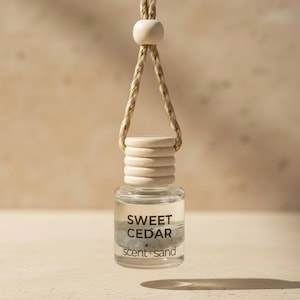 May include: A small, clear glass bottle filled with liquid and sand, featuring a light-colored wooden cap and a hanging cord with a wooden bead. The bottle is printed with the text SWEET CEDAR scent sand in black lettering on the front surface.