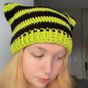 May include: A crochet beanie featuring a cat-ear silhouette with horizontal stripes in alternating neon lime green and black yarn. The hat is constructed with a ribbed brim and a textured stitch pattern throughout, creating two distinct pointed ears at the top.