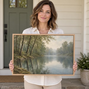 May include: A rectangular landscape painting in a light wood frame depicting a calm river flowing through a forest with birch trees on the bank. The scene features soft, muted green and blue tones with a hazy, overcast sky reflecting on the water.