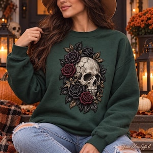 May include: A dark forest green crewneck sweatshirt featuring a central graphic of a cracked human skull surrounded by a cluster of deep red and black roses with dark green leaves. The design is detailed with intricate shading and textures.