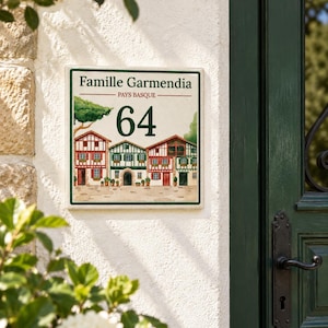 Puede incluir: Un letrero cuadrado de cerámica para casa con el texto Famille Garmendia y Pays Basque sobre el número 64 en verde oscuro. El diseño incluye una colorida ilustración de tres casas de entramado de madera con detalles rojos y verdes.