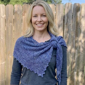 May include: A triangular knit shawl in a variegated blue yarn with a loose, open-stitch texture. The shawl features a pointed bottom edge and is tied in a knot at the side, layered over a dark grey long-sleeved shirt with a simple crew neckline.