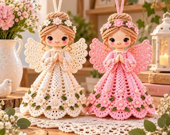 May include: Two crocheted angel ornaments with light brown yarn hair, floral crowns, and large wings. One angel wears a white dress with flower accents, and the other wears a pink dress with flower accents. Both have hands in a prayer position.