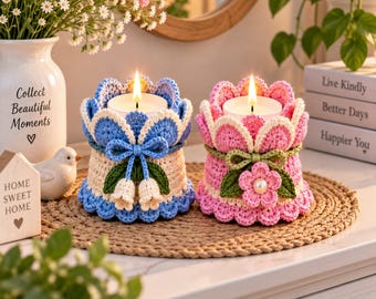 May include: Two cylindrical candle holders with crocheted covers. One cover is light blue and cream with a blue bow and two small white flowers. The other is pink and cream with a green bow and a pink flower with a central pearl bead.