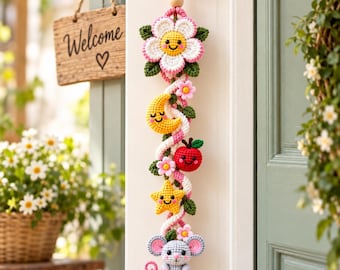 May include: A vertical crochet wall hanging featuring a series of colorful, smiling figures including a white flower, a yellow crescent moon, a red apple, a yellow star, and a gray mouse, all connected by a twisted pink and white cord.
