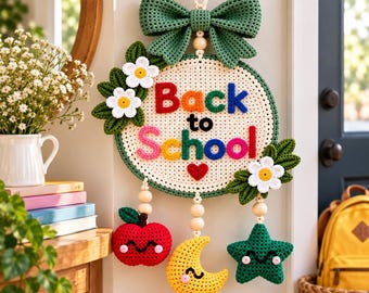 May include: A round crochet wall hanging featuring the text Back to School in colorful yarn letters. It is decorated with a large green bow, white flowers, and three dangling crochet charms: a red apple, a yellow crescent moon, and a green star.