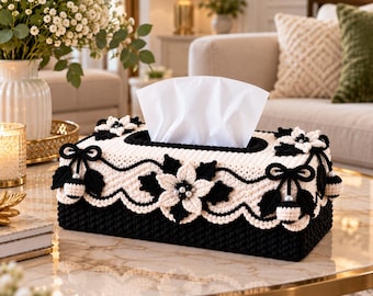 May include: A rectangular tissue box cover featuring a black base and a cream-colored top. The cover is decorated with crocheted floral motifs, black and cream bows, and small white beads. A stack of white tissues protrudes from the center opening.
