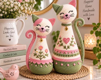 May include: Two handmade crochet cat figurines in cream, sage green, and soft pink. The smaller cat features a floral design, while the larger cat has a heart motif. Both cats have curled tails, embroidered faces, and small gold-toned heart charms.