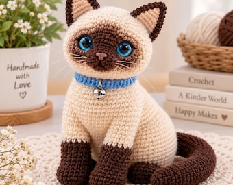 May include: A crocheted Siamese cat plush toy with cream-colored fur, dark brown ears, face, and paws, and bright blue eyes. The cat wears a light blue collar with a small silver bell attached. The yarn texture is visible throughout the entire figure.