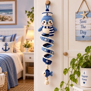 May include: A crocheted hanging decoration featuring a blue and white seal wearing a sailor hat and scarf, attached to a long, spiraling blue and white ribbon adorned with small white star accents, ending with a small striped fish and a tassel.