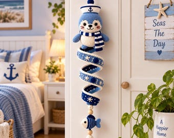 May include: A crocheted hanging decoration featuring a blue and white seal wearing a sailor hat and scarf, attached to a long, spiraling blue and white ribbon adorned with small white star accents, ending with a small striped fish and a tassel.