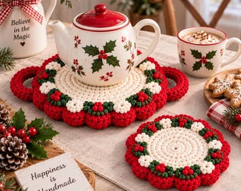 May include: A cream-colored ceramic teapot with a red lid and holly berry pattern, resting on a round, red, white, and green crocheted trivet with handles. A matching smaller crocheted coaster and a mug with a holly pattern are nearby.
