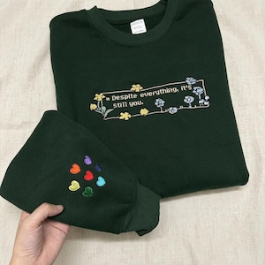 Peut inclure: Un sweat-shirt à col rond vert forêt foncé avec un cadre rectangulaire brodé contenant le texte "Despite everything, it's still you." entouré de petits motifs floraux. La manche présente un groupe de sept petits cœurs multicolores.