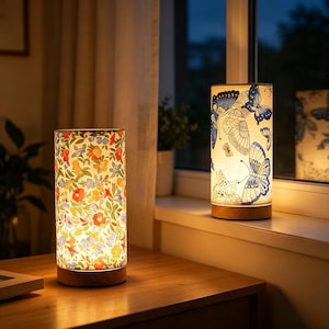 Peut inclure: Deux lampes de table cylindriques avec des bases en bois. Une lampe présente un motif floral multicolore vif avec des fleurs rouges, orange, jaunes et bleues. L'autre lampe affiche un imprimé papillon bleu et blanc sur fond clair.