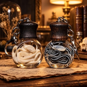 May include: Two spherical glass bottle keychains with dark wooden caps. The left bottle contains a coiled off-white snake, while the right bottle holds a coiled black and white patterned snake. Both feature silver metal key rings and chains.