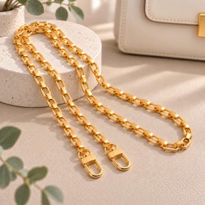 May include: A gold-toned metal chain strap featuring a series of interconnected rectangular links. The strap is finished with two matching gold-toned lobster claw clasps at each end, designed for attachment to a handbag or similar accessory.