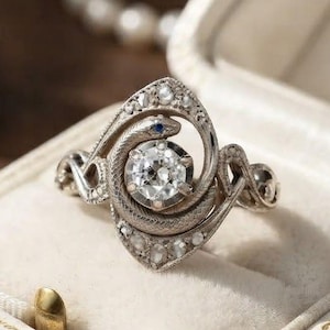 Op de afbeelding: Een zilverkleurige ring met een ontwerp van een kronkelende slang die een centrale, rondgeslepen, heldere edelsteen omsluit. De slang heeft een klein blauw accentsteentje als oog en de ringband is versierd met kleine heldere steentjes.