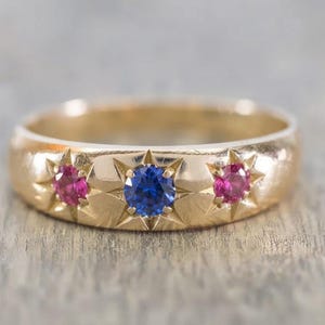 May include: A gold band ring featuring three round-cut gemstones set in star-shaped engravings. The centre stone is a vibrant blue sapphire, flanked by two smaller, deep pink rubies. The metal surface has a polished finish with starburst details.