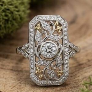 May include: A rectangular silver-toned ring featuring a central round-cut stone surrounded by intricate filigree leaf patterns and small gold-toned accents. The entire outer border and the band are encrusted with numerous tiny, sparkling white stones.