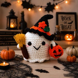 May include: A white crocheted ghost figure wearing a black and orange witch hat with a small orange star and green accent. The ghost holds a brown and yellow broom in one hand and a small orange crocheted jack-o'-lantern bucket in the other.