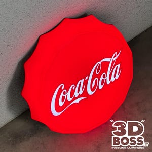 Può includere: Un'insegna luminosa circolare di colore rosso brillante a forma di tappo a corona, che riporta il logo Coca-Cola in bianco con il caratteristico carattere corsivo. L'insegna emette una luce vivace e include il testo 3D BOSS.IT in basso a destra.