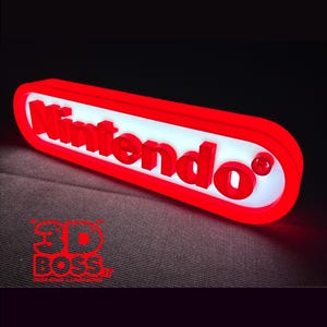 以下が含まれることがあります： 赤と白のNintendoのイルミネーション看板。白い楕円形の背景に太い赤の3D文字でブランド名が記され、厚い赤い枠で囲まれています。看板のテキストの横には登録商標マークが表示されています。