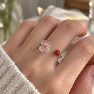 May include: An open-style ring featuring a translucent, pale pink flower with a small coral-colored center bead on one end and a single, smooth, solid red spherical bead on the other end, set on a clear, thin, flexible band.