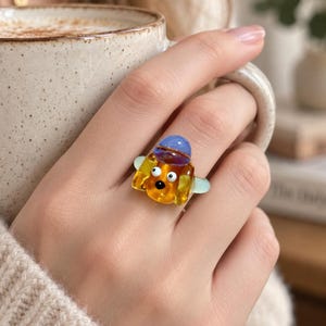 May include: A glass ring featuring a small, amber-colored dog figure with white eyes and a black nose. The dog wears a blue hat on its head and has light green ears. The ring band is a translucent light green color, creating a playful accessory.