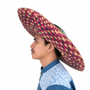 May include: A wide-brimmed woven hat featuring a geometric pattern in shades of magenta, dark green, and natural tan. The hat has a high crown and a broad, flat brim. It is worn with a blue shirt that has a light blue patterned shoulder panel.