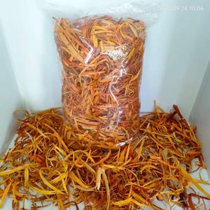 Op de afbeelding: Een doorzichtige plastic zak gevuld met dunne, gedroogde, oranje en gele houtsnippers of baststroken. Een hoop van hetzelfde losse, vezelige, goudoranje houtmateriaal is voor de zak uitgespreid, wat een getextureerd en versnipperd uiterlijk geeft.