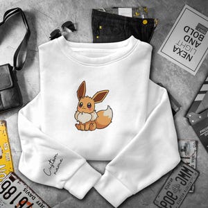 May include: A white long-sleeve sweatshirt featuring a centered graphic of a brown Eevee character with large eyes and a fluffy cream-colored chest. The sleeve includes the black cursive text Custom name printed near the wrist cuff.