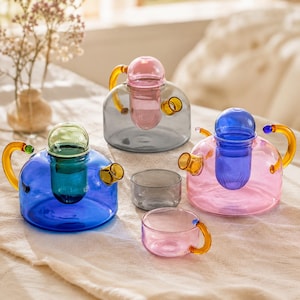 Puede incluir: Dos teteras de cristal y dos tazas pequeñas de cristal. Una tetera es azul con tapa verde y asa ámbar, la otra es gris con tapa rosa y asa ámbar. Una tetera rosa con tapa azul y asa ámbar, además de dos tazas pequeñas de cristal.
