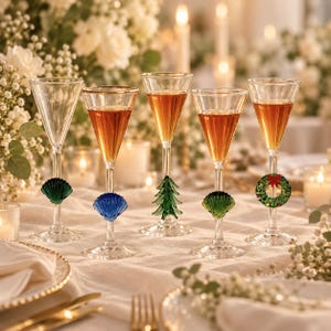 May include: Five clear glass stemmed glasses with decorative charms on the stems. The charms include a green seashell, a blue seashell, a green Christmas tree, a green seashell, and a green Christmas wreath with a red bow. Four glasses contain amber liquid.