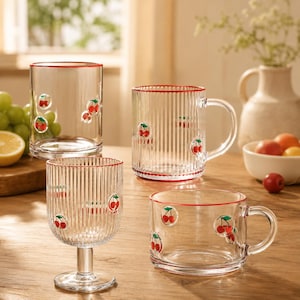 May include: A collection of four clear glass vessels featuring red cherry illustrations and thin red rims. The set includes a tall tumbler, a ribbed mug, a ribbed stemmed glass, and a wide-mouthed mug, all decorated with small red cherry motifs.