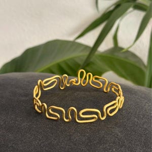Può includere: Un bracciale color oro realizzato con un unico filo continuo piegato in una serie ripetitiva di anelli rettangolari arrotondati e interconnessi che formano una fascia circolare con un piccolo meccanismo di chiusura visibile in alto.