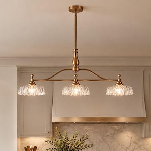 May include: A linear chandelier featuring a brass-toned metal frame with a central rod and three downward-facing lights. Each light is encased in a scalloped, clear glass shade with a textured, ribbed pattern that diffuses the light.