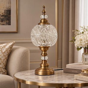 Puede incluir: Una lámpara de mesa con una pantalla redonda de vidrio transparente con patrón de diamante, apoyada sobre una base de metal con acabado en latón antiguo. La lámpara incluye un remate decorativo en la parte superior y un cristal en el eje.