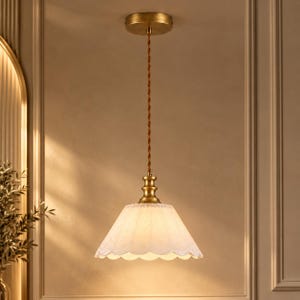 Puede incluir: Una lámpara colgante con florón de techo y portalámparas en tono latón. Un cable trenzado marrón sostiene una pantalla de cristal blanco translúcido con bordes festoneados que emite una luz suave y cálida hacia abajo.