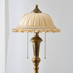 Puede incluir: Una l&aacute;mpara de mesa con una pantalla de cristal &aacute;mbar acanalada en forma de c&uacute;pula con borde festoneado. La base es de metal en tono lat&oacute;n antiguo con detalles ornamentados y dos cadenas de tiro que cuelgan bajo la pantalla.