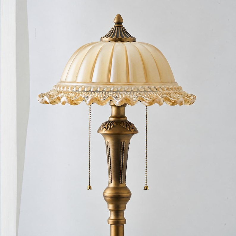 Puede incluir: Una l&aacute;mpara de mesa con una pantalla de cristal &aacute;mbar acanalada en forma de c&uacute;pula con borde festoneado. La base es de metal en tono lat&oacute;n antiguo con detalles ornamentados y dos cadenas de tiro que cuelgan bajo la pantalla.