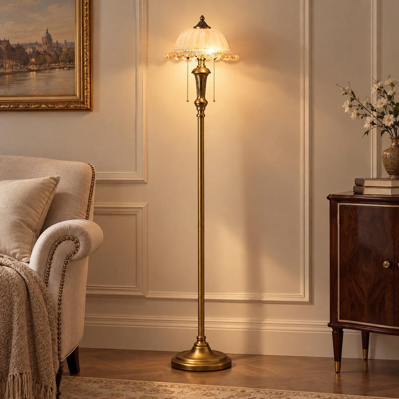 May include: A tall floor lamp with a metallic brass-toned finish, featuring a slender central pole and a circular base. The lamp is topped with a white, fluted glass shade adorned with hanging crystal-like accents and two pull chains.