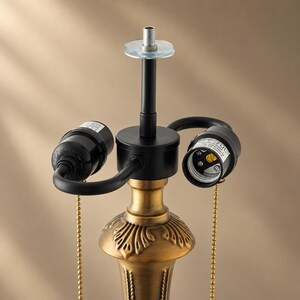 Puede incluir: Una base de l&aacute;mpara con acabado en lat&oacute;n cepillado y detalles de hojas ornamentadas en el cuello. Incluye dos casquillos negros para bombillas, cada uno con una cadena de tracci&oacute;n dorada, y un v&aacute;stago central de metal negro con un remate.