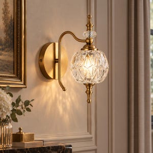 Puede incluir: Un aplique de pared con acabado en latón cepillado, un brazo metálico curvado y una pantalla esférica de cristal transparente facetado. El diseño incluye un remate decorativo en la parte superior e inferior, con un pequeño cristal.