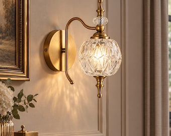 May include: A wall-mounted light fixture featuring a brushed brass finish, a curved metal arm, and a spherical, faceted clear glass shade. The design includes a decorative finial at the top and bottom, with a small crystal accent piece.