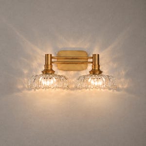 Puede incluir: Un aplique de pared doble con una placa trasera de acabado en latón cepillado y dos brazos horizontales que sostienen dos pantallas de vidrio transparente festoneado orientadas hacia abajo, emitiendo una luz cálida con patrones.