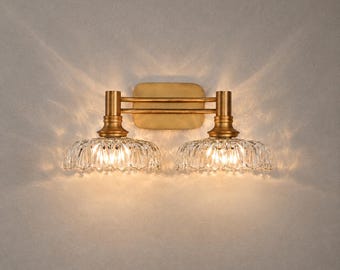 May include: A double wall sconce featuring a brushed brass finish backplate and two horizontal arms supporting two downward-facing, scalloped clear glass shades that emit a warm, glowing light with distinct light patterns on the wall.