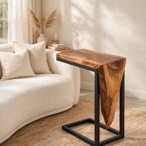 May include: A C-shaped side table featuring a natural live-edge wood top and side panel with a visible grain pattern, supported by a black rectangular metal frame base. The table has a smooth, polished finish on the horizontal surface.